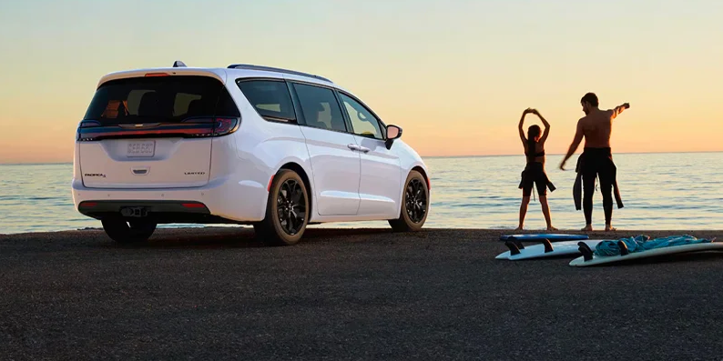 A photo of a white 2025 Chrysler Pacifica parker on the beach with 2 people and their surf boards next to them.